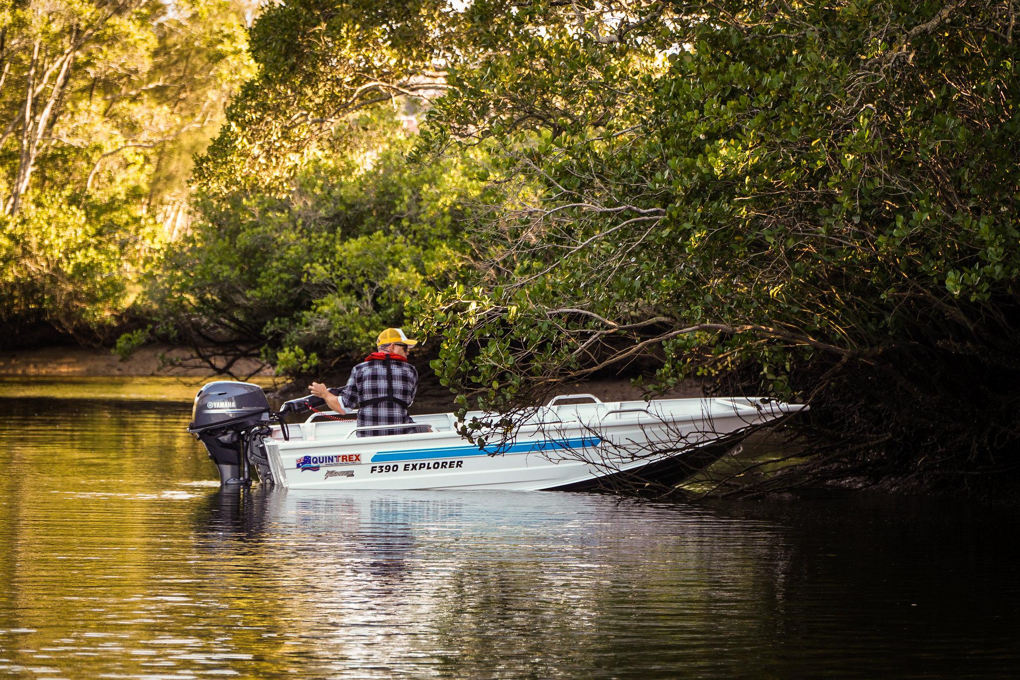 A man operating an outboard engine of a small boat on a lake's shore.