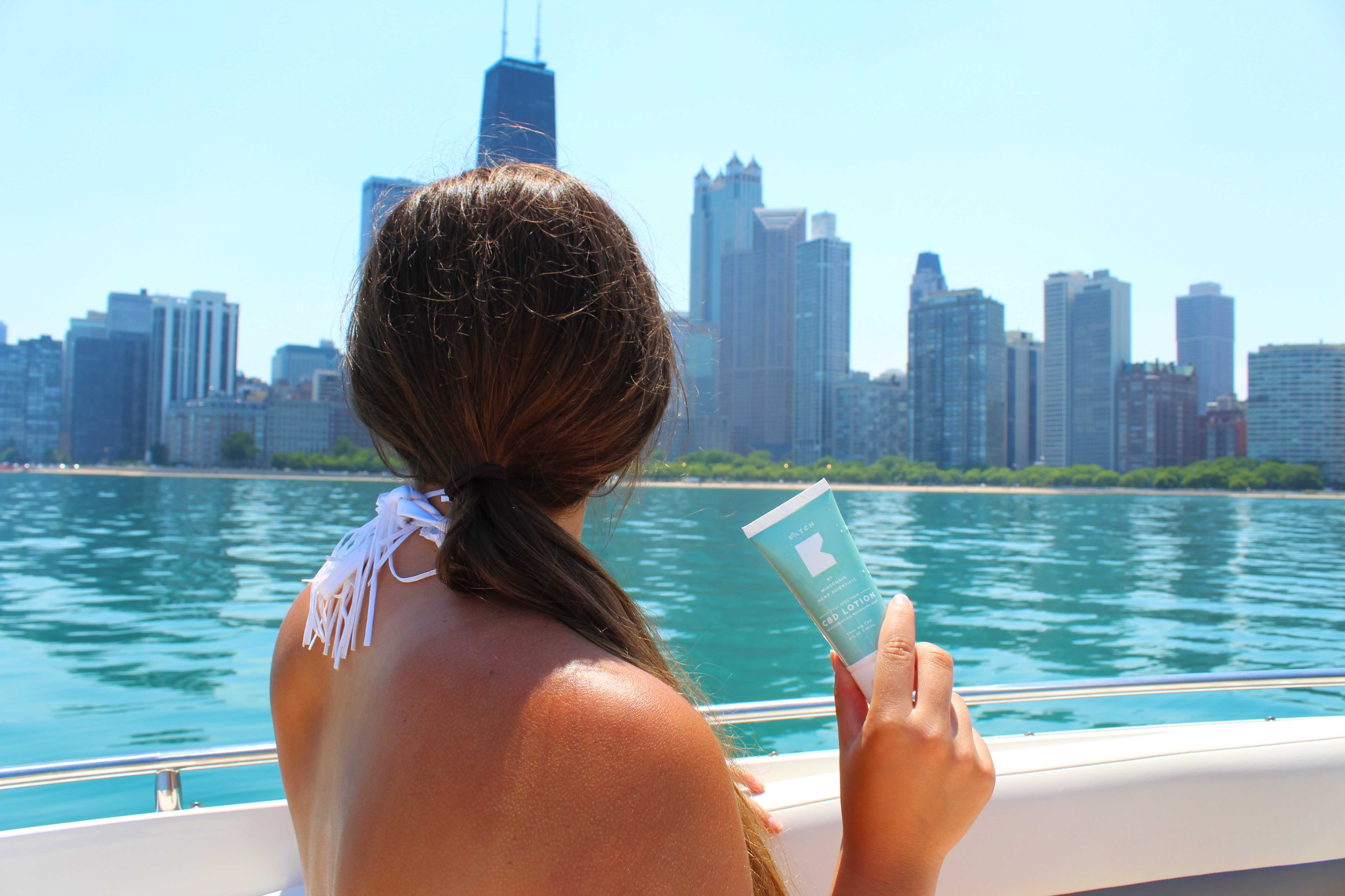 A woman sitting on a boat is facing is her back at the camera and holding a sunscreen.