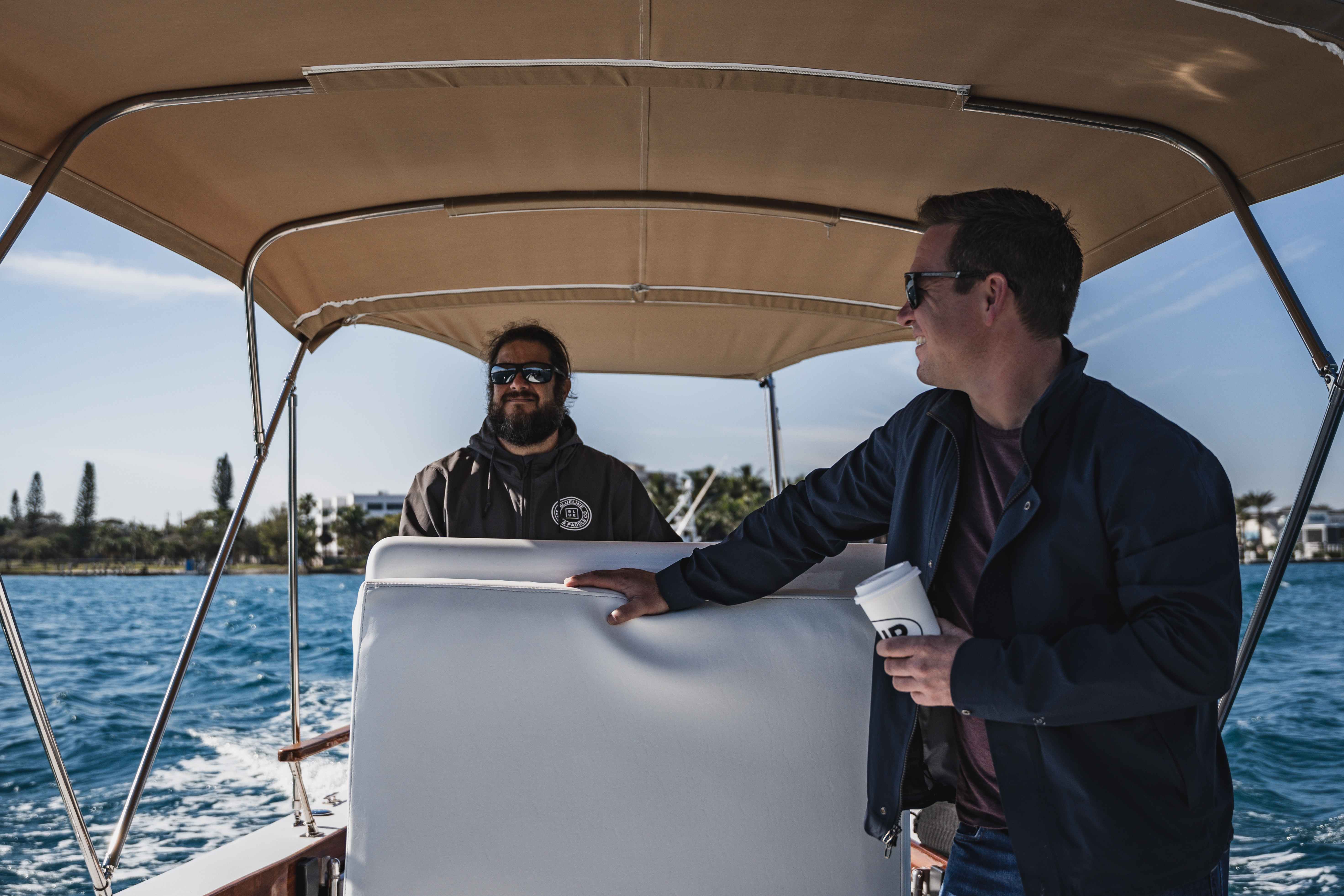 A man wearing shades driving a motor boat  with a canopy while another man is smiling at him.