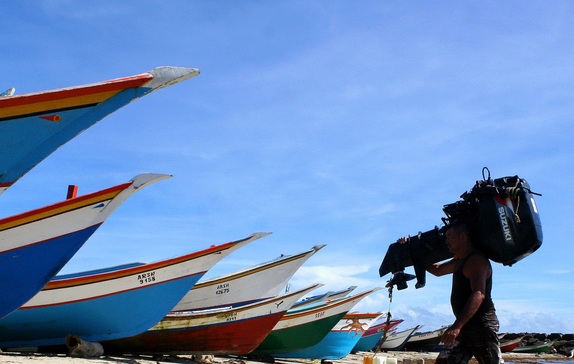 A man carrying a Suzuki outboard with a row of boats in the background.