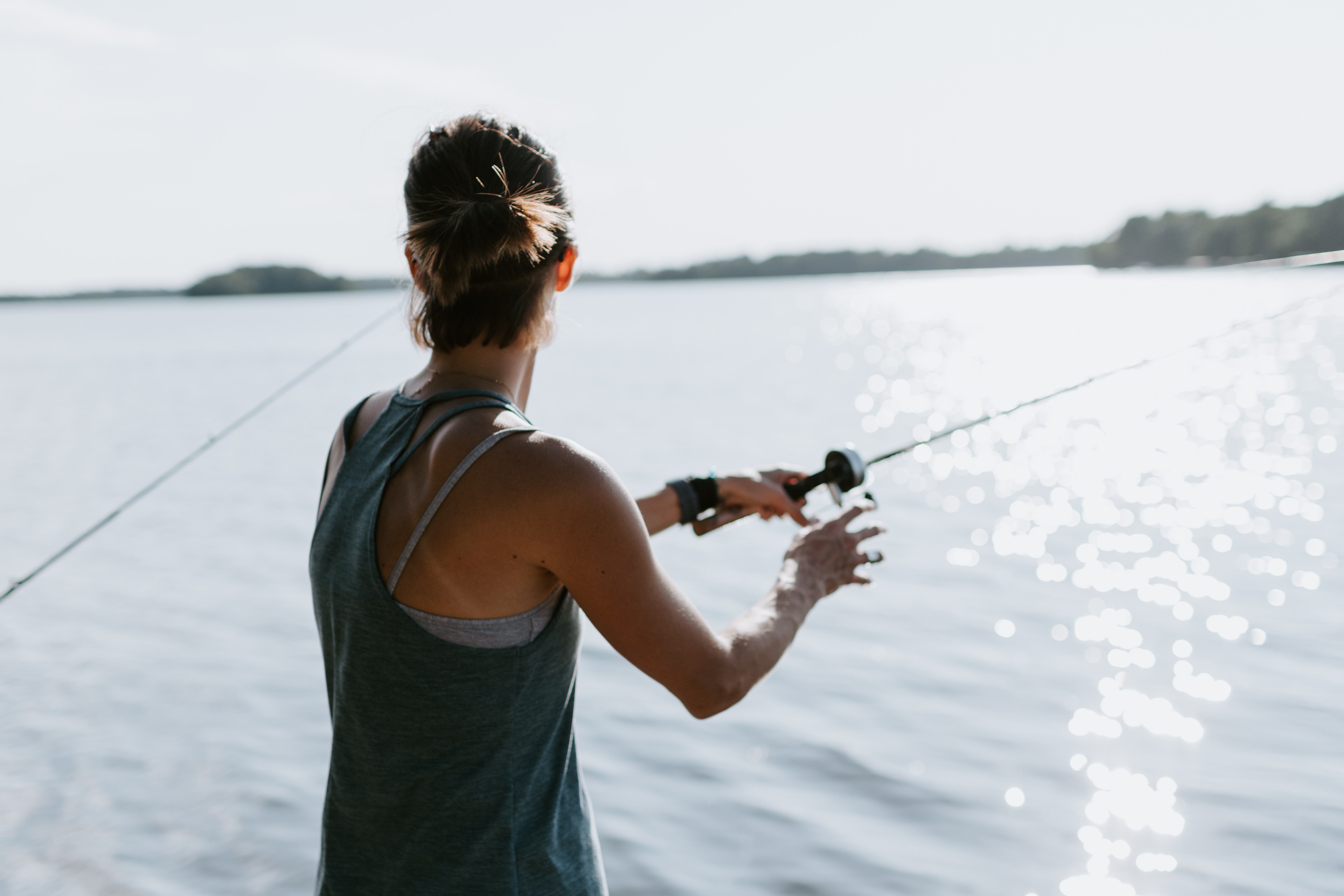 A woman fly-fishing at a lake while wearing a sleeveless top.