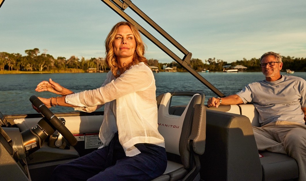 A woman wearing a pink cap, purple shirt, shorts, and brown boots is sitting at the bow of a boat