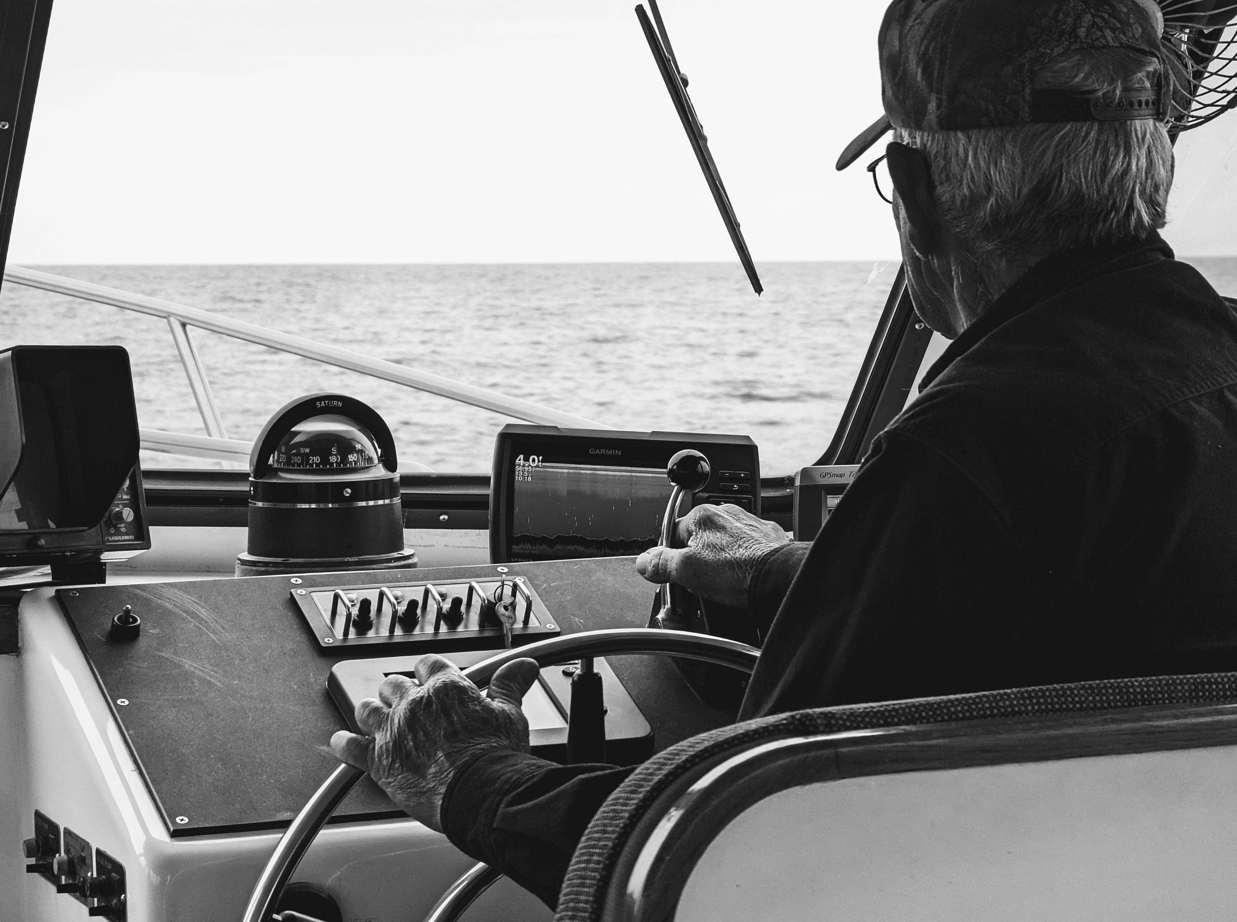 A black and white photo of an old man wearing a cap operating a boat's console and steering wheel