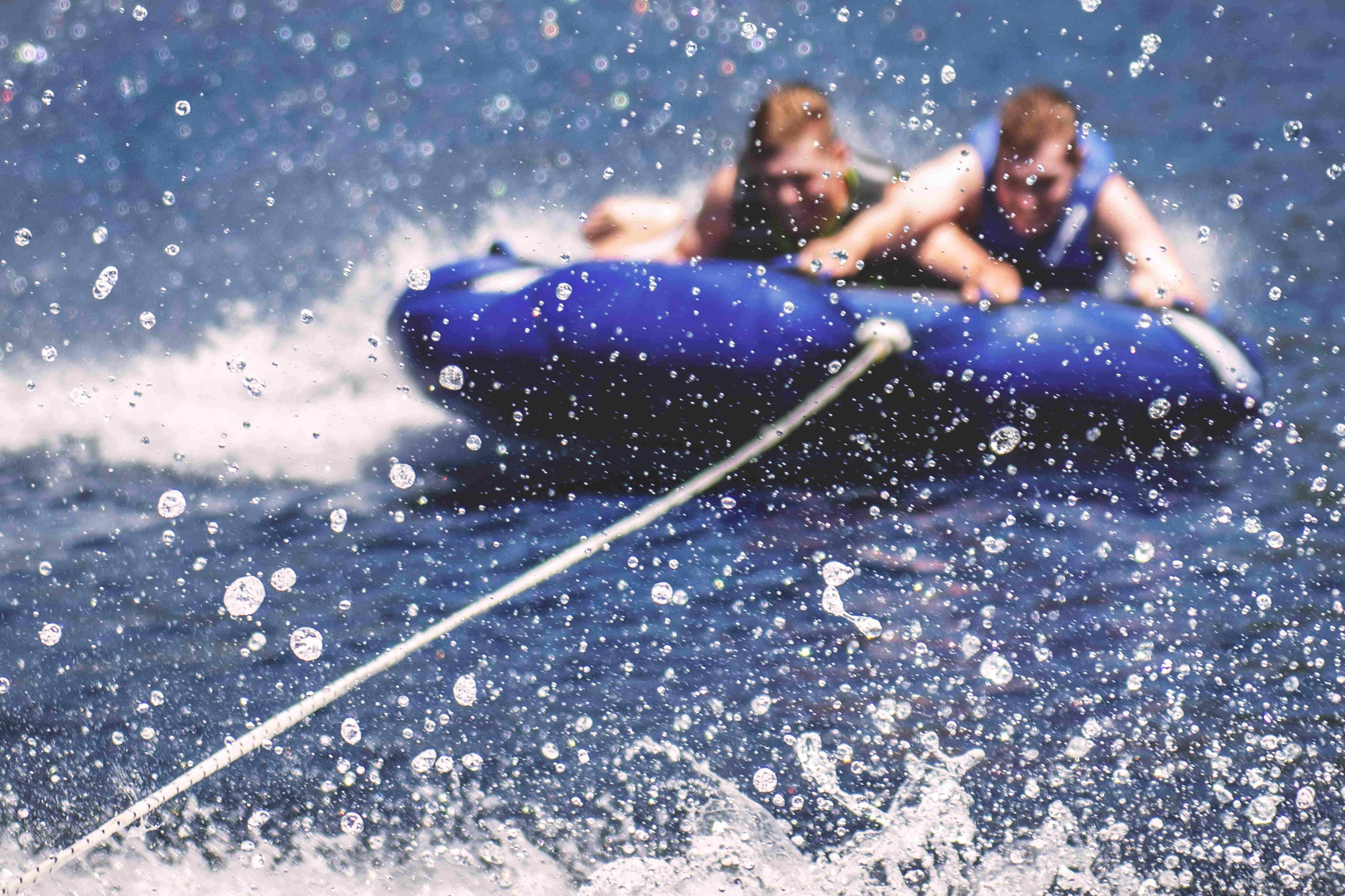 Two men on a tube while being towed behind a boat making a huge splash.